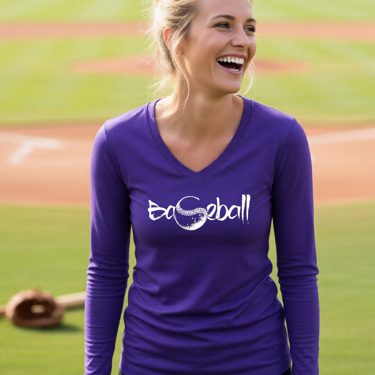 A woman smiling in front of a baseball diamond wearing a purple long-sleeve t-shirt, with an artistic white graphic of a sketch of the word BASEBALL” in a curved font with the S replaced by a baseball, printed on the front of the shirt.