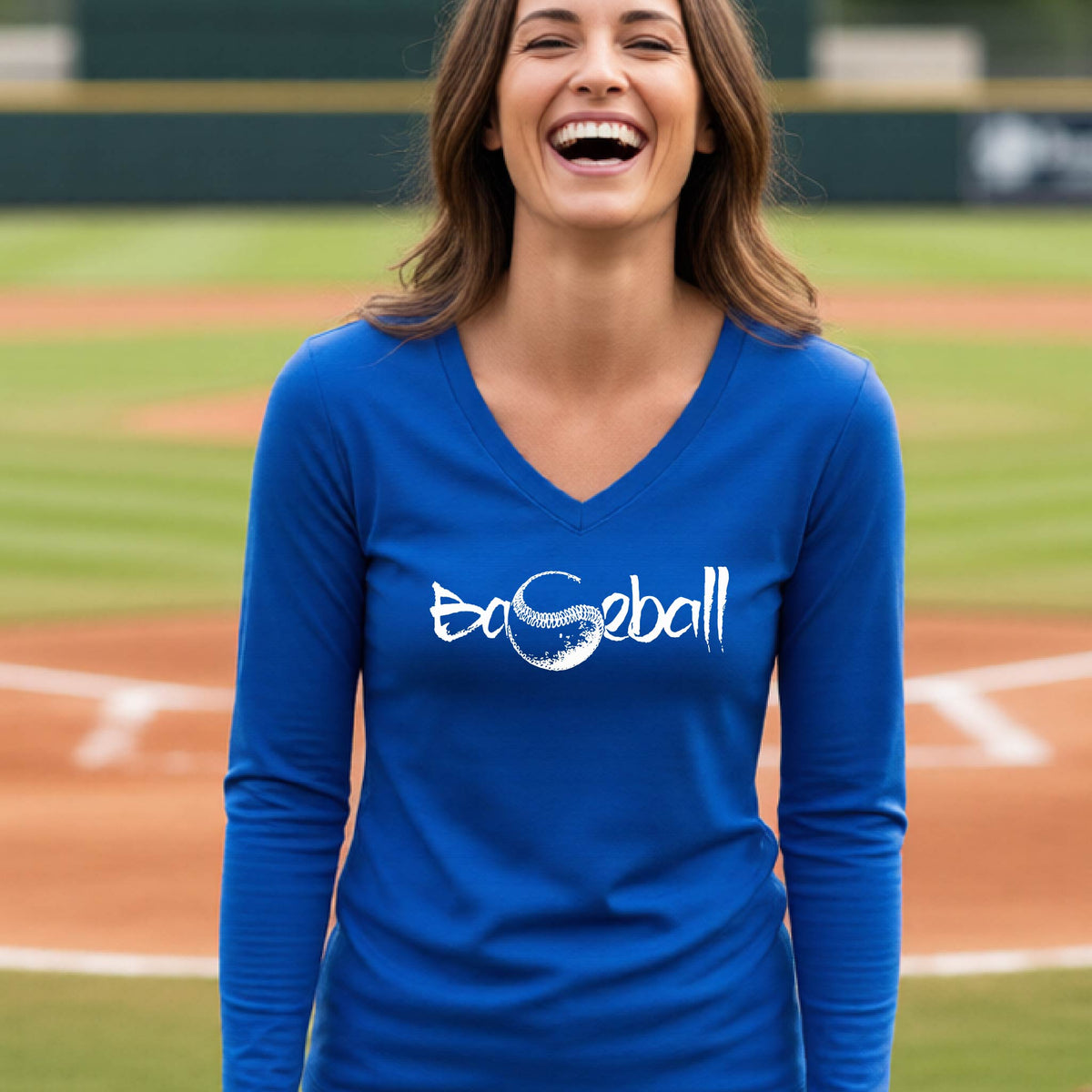 A woman smiling in front of a baseball diamond wearing a blue long-sleeve t-shirt, with an artistic white graphic of a sketch of the word BASEBALL” in a curved font with the S replaced by a baseball, printed on the front of the shirt.