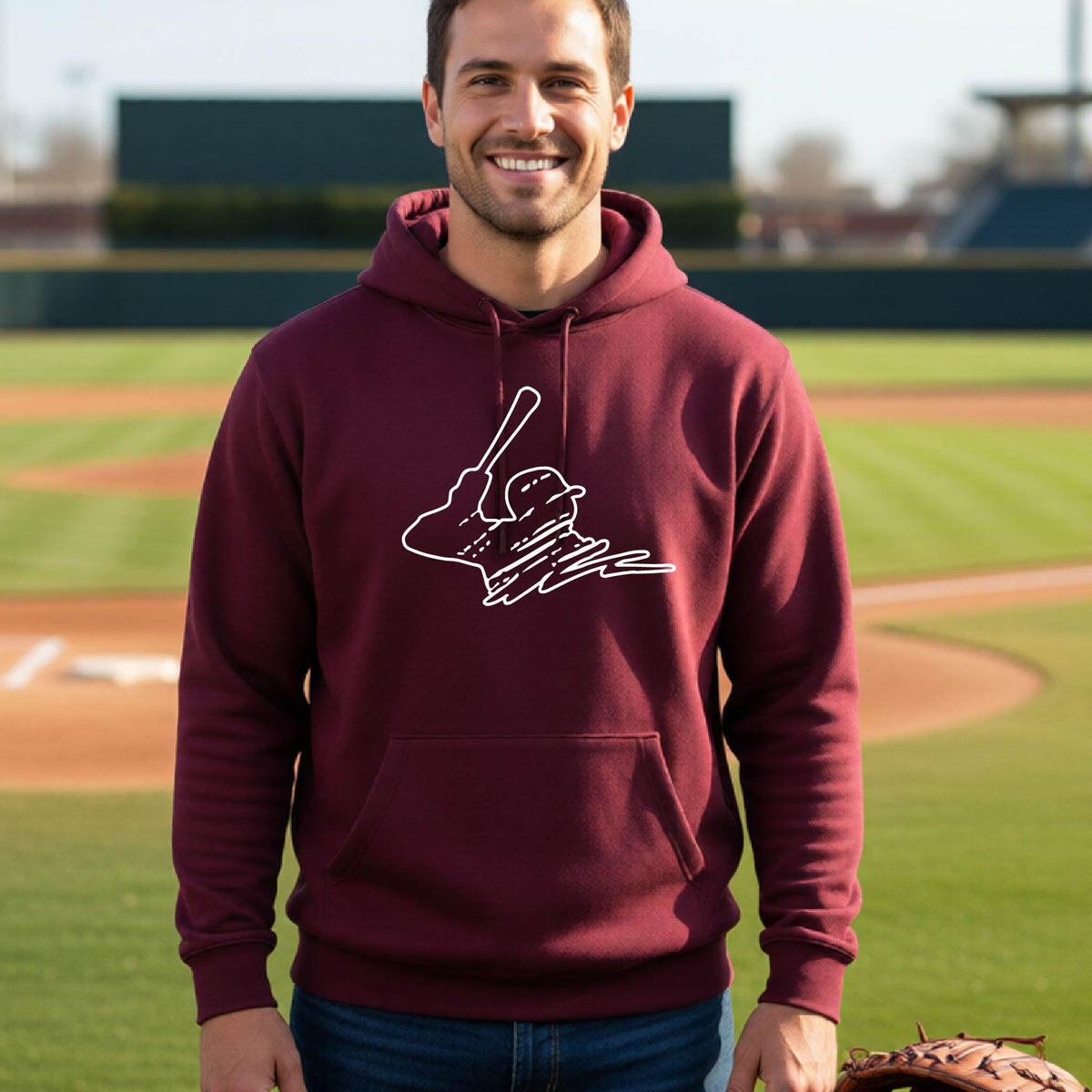 A man smiling in front of a baseball diamond wearing a maroon hoodie pullover, with a white graphic of an outline of a baseball player up to bat, printed on the front.