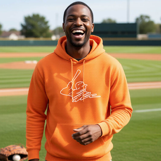 A man smiling in front of a baseball diamond wearing an orange hoodie pullover, with a white graphic of an outline of a baseball player up to bat, printed on the front.