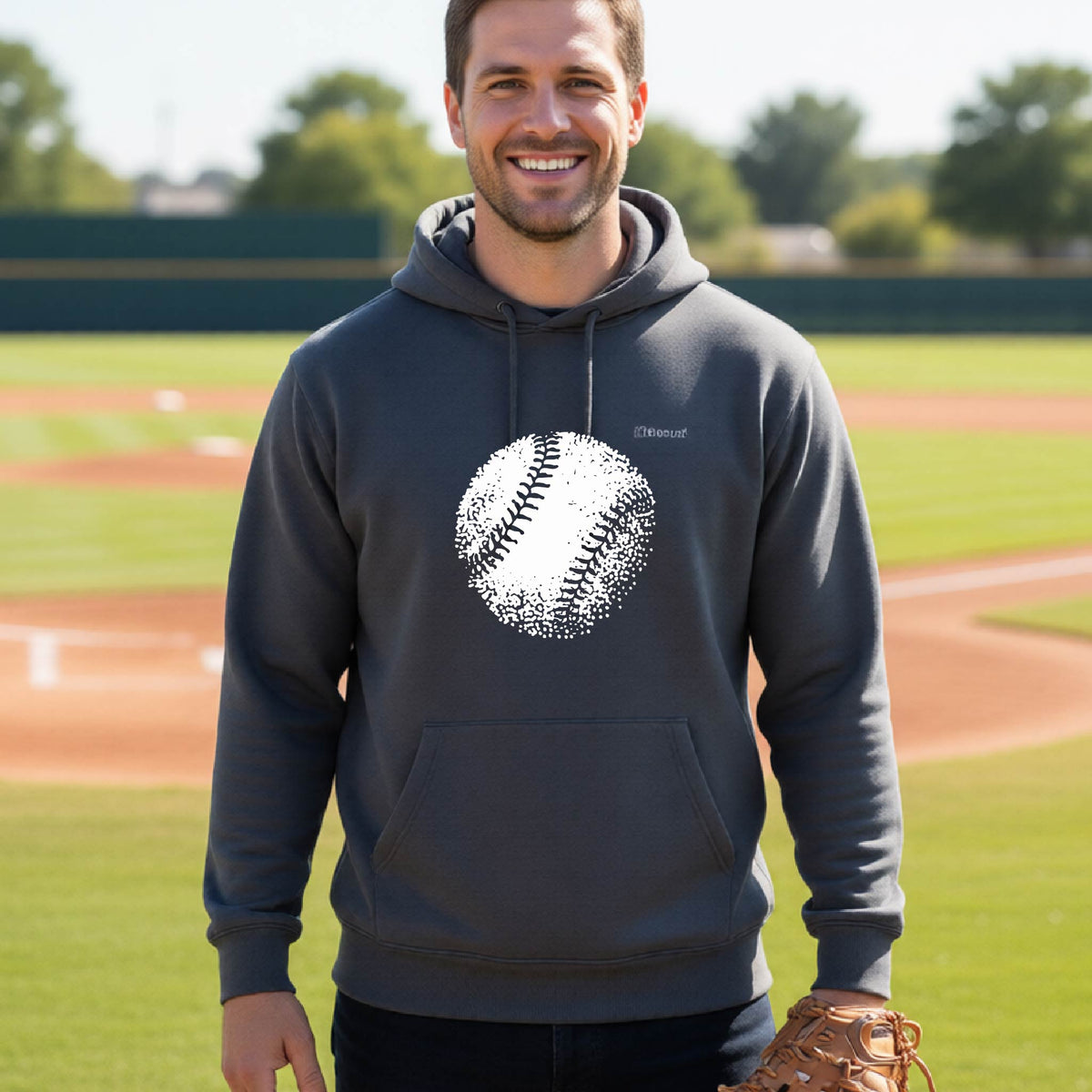 A man smiling in front of a baseball diamond wearing a dark gray pullover hoodie, with an artistic white graphic of a retro looking baseball, printed on the front of the shirt.