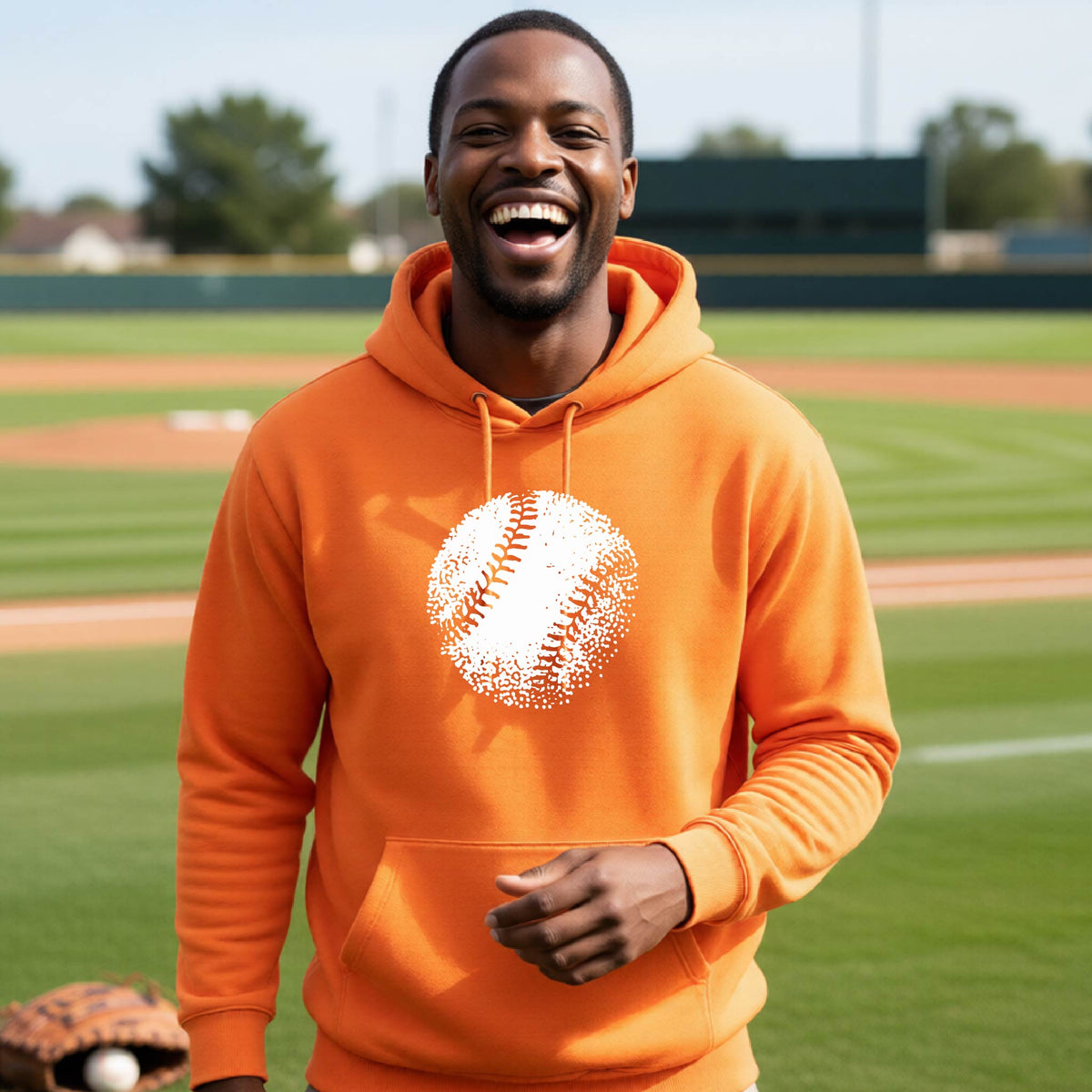A man smiling in front of a baseball diamond wearing an orange pullover hoodie, with an artistic white graphic of a retro looking baseball, printed on the front of the shirt.