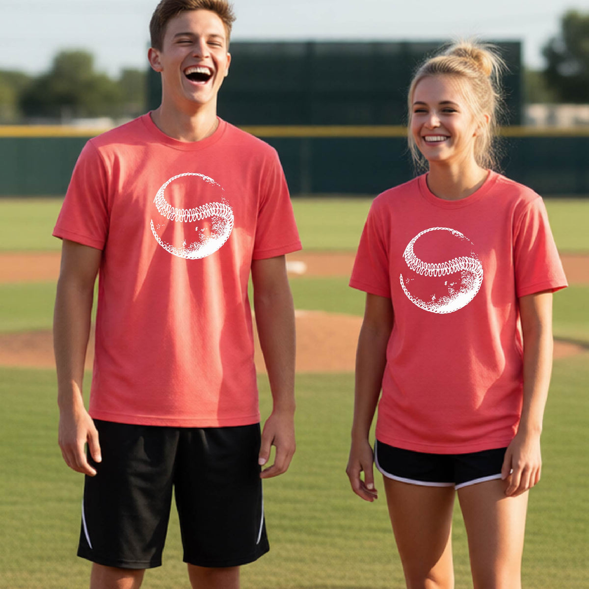 Two youth athletes wearing matching shirts, smiling in front of a baseball diamond wearing coral colored short-sleeve t-shirts, with a white artistic graphic of a baseball, printed on the front.