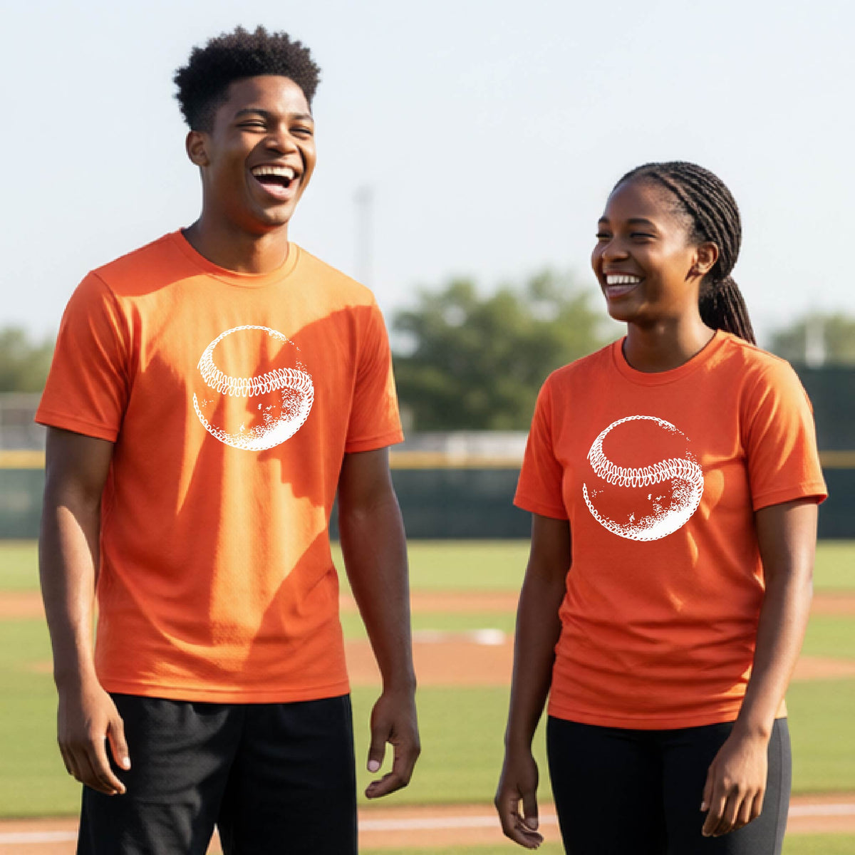 Two youth athletes wearing matching shirts, smiling in front of a baseball diamond wearing orange short-sleeve t-shirts, with a white artistic graphic of a baseball, printed on the front.