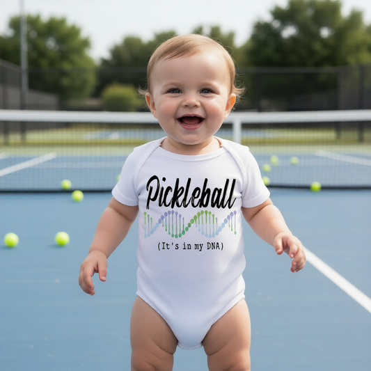 A smiling baby standing on a blue pickleball court wearing a white bodysuit with the text 'Pickleball (It's in my DNA)' and a small multicolor graphic.