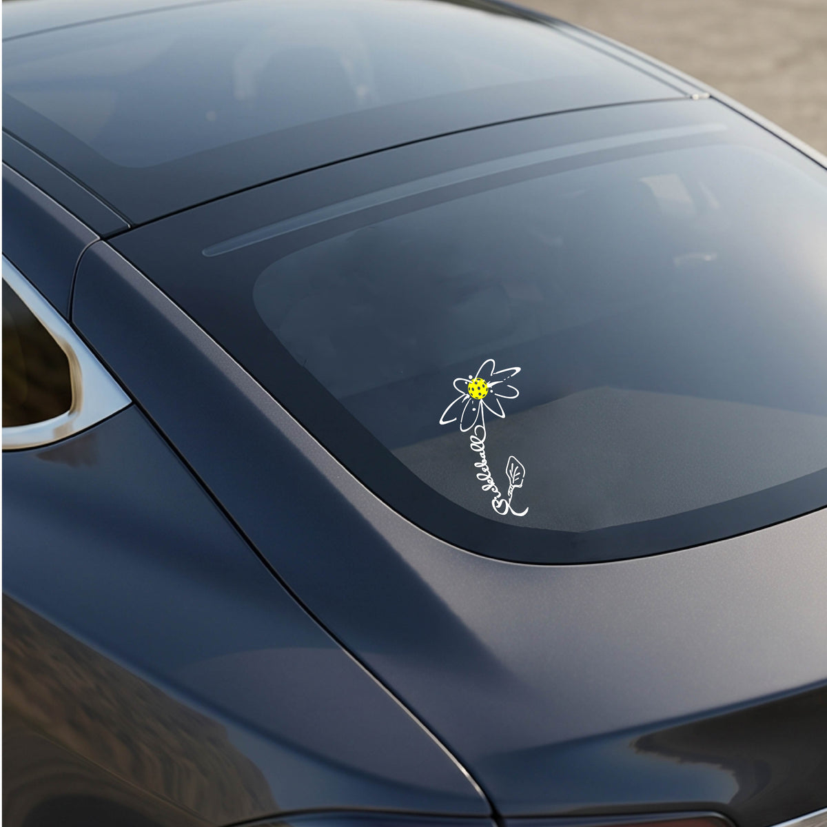 A car window with a white pickleball flower with a stem made out of the text 'PICKLEBALL' and the leaf a pickleblal paddle.applied to the glass.