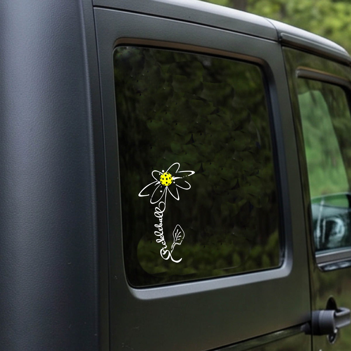 A car window with a white pickleball flower with a stem made out of the text 'PICKLEBALL' and the leaf a pickleblal paddle.applied to the glass.