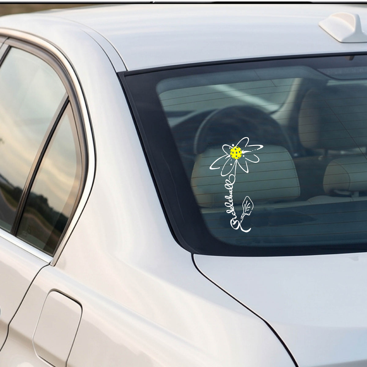 A car window with a white pickleball flower with a stem made out of the text 'PICKLEBALL' and the leaf a pickleblal paddle.applied to the glass.