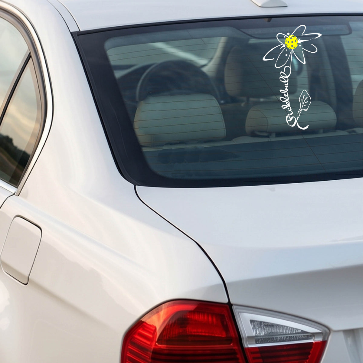 A car window with a white pickleball flower with a stem made out of the text 'PICKLEBALL' and the leaf a pickleblal paddle.applied to the glass.