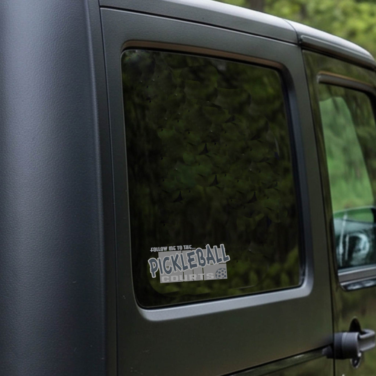 A car window showing a 'FOLLOW ME TO THE PICKLEBALL COURTS' vinyl decal sticker applied to a vehicle window. 