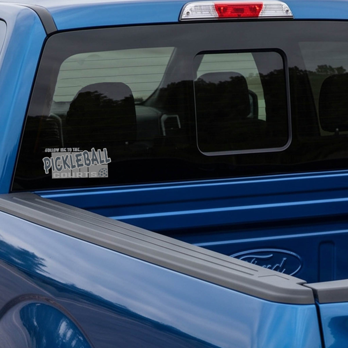 A car window showing a 'FOLLOW ME TO THE PICKLEBALL COURTS' vinyl decal sticker applied to a vehicle window. 