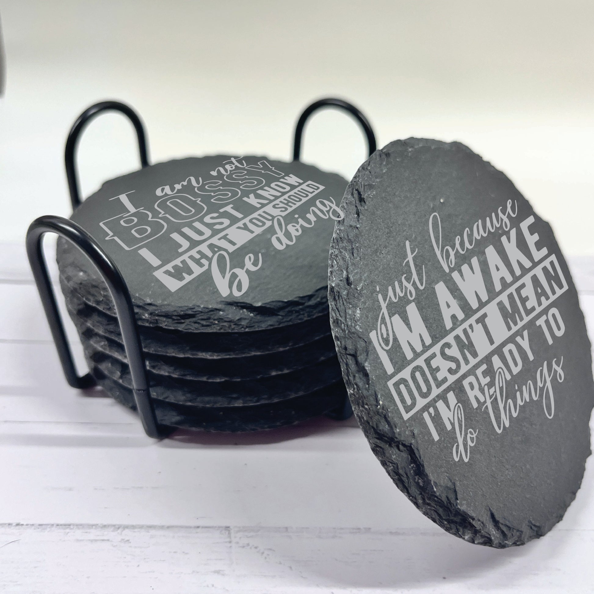 Set of slate coasters with humorous sayings in a black holder on a light wooden surface.