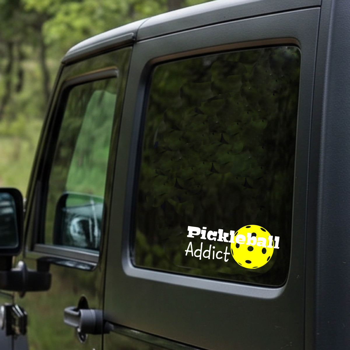 A black car rear window with a 'Pickleball Addict' decal featuring a yellow pickleball, shown in a real-world outdoor setting.
