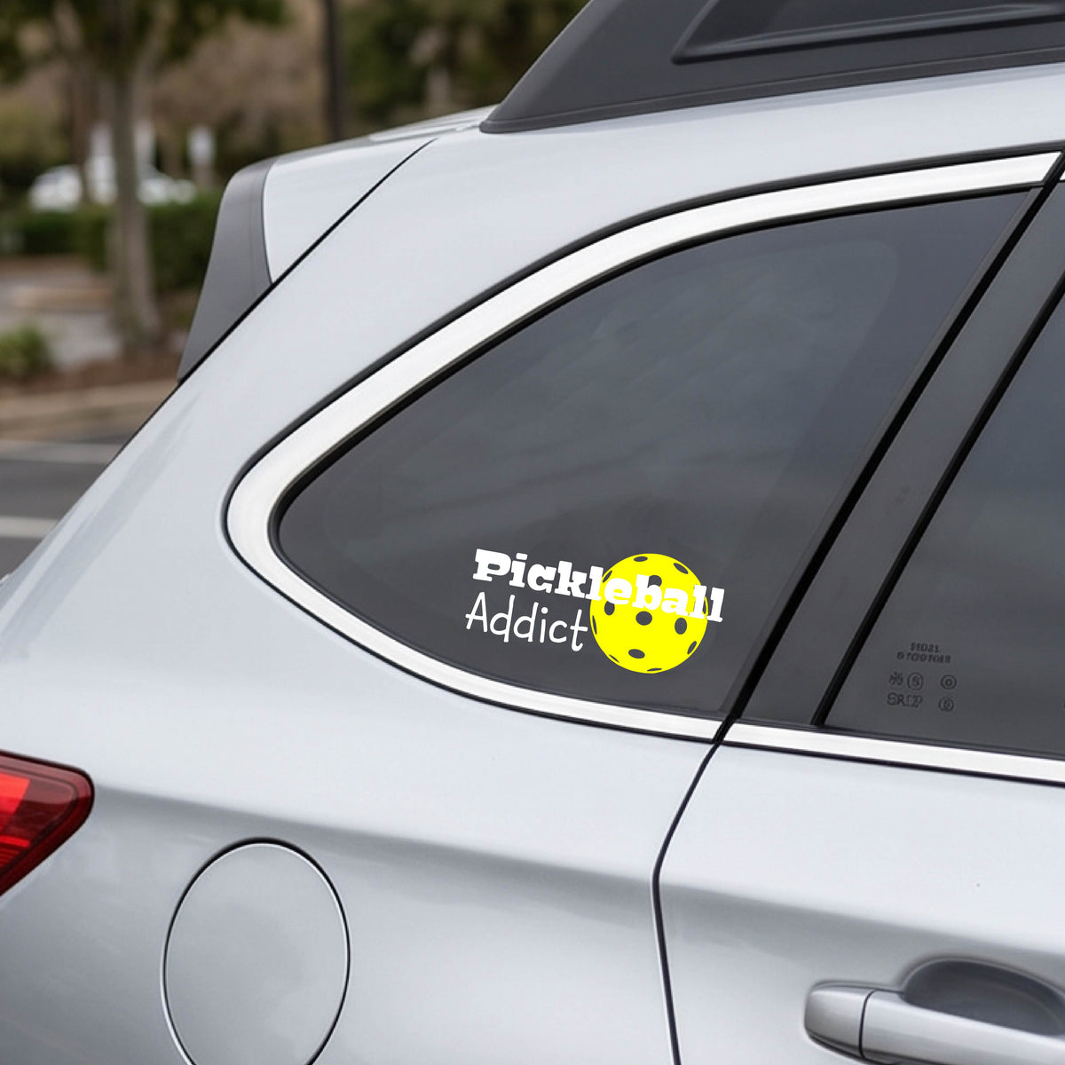 A silver car rear window with a 'Pickleball Addict' decal featuring a yellow pickleball, shown in a real-world outdoor setting.