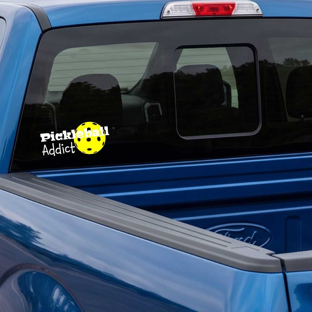 A blue truck rear window with a 'Pickleball Addict' decal featuring a yellow pickleball, shown in a real-world outdoor setting.