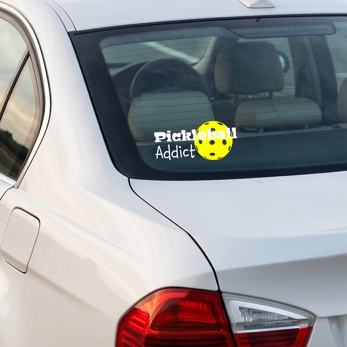 A white car rear window with a 'Pickleball Addict' decal featuring a yellow pickleball, shown in a real-world outdoor setting.