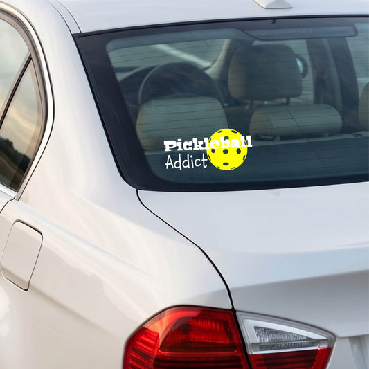 A white car rear window with a 'Pickleball Addict' decal featuring a yellow pickleball, shown in a real-world outdoor setting.