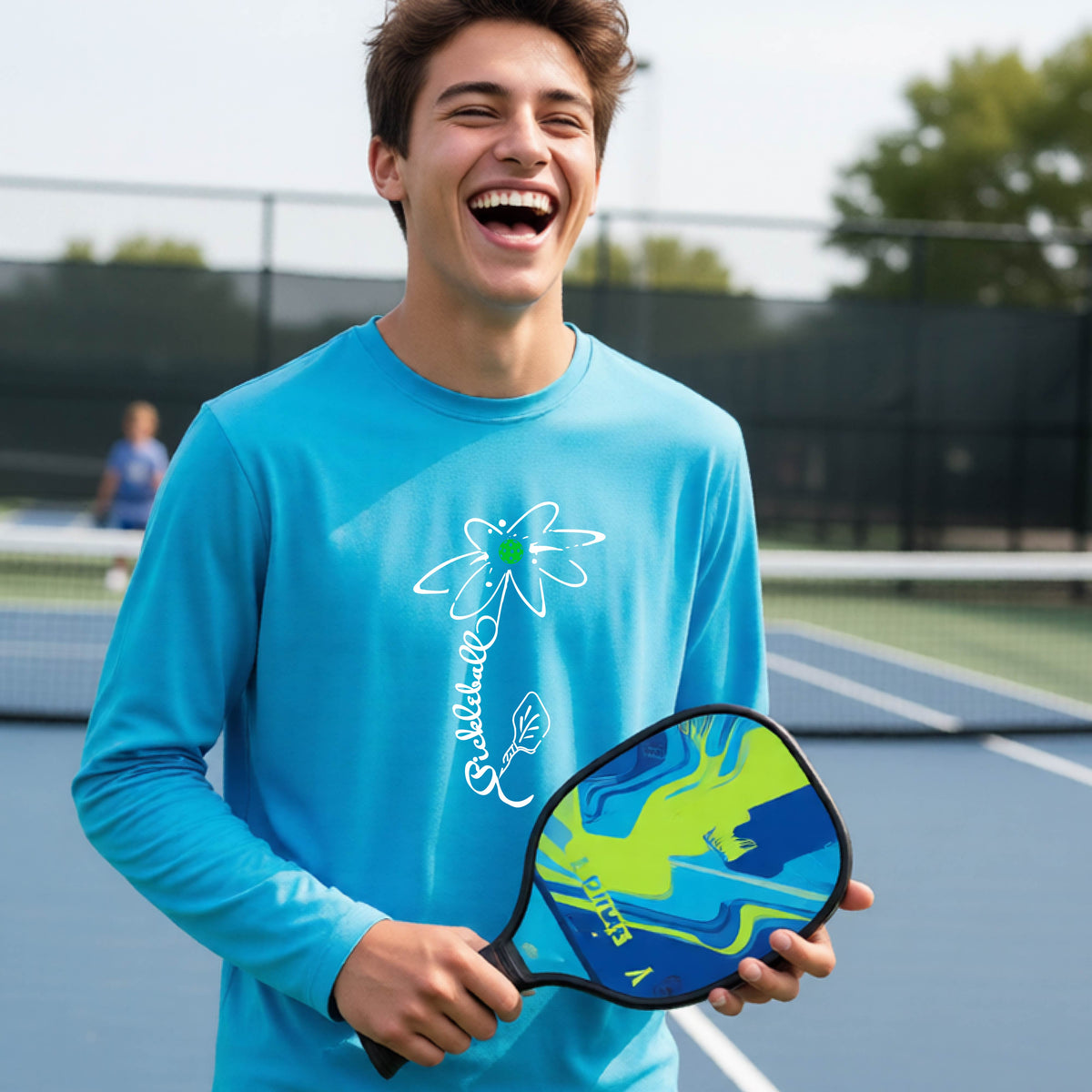 A youth wearing a bright blue long-sleeve t-shirt with a pickleball graphic of a flower with a colorful pickleball as the center with  the stem made of the word ‘PICKLEBALL’ and the leaf is a pickleball paddle, standing on a pickleball court.
