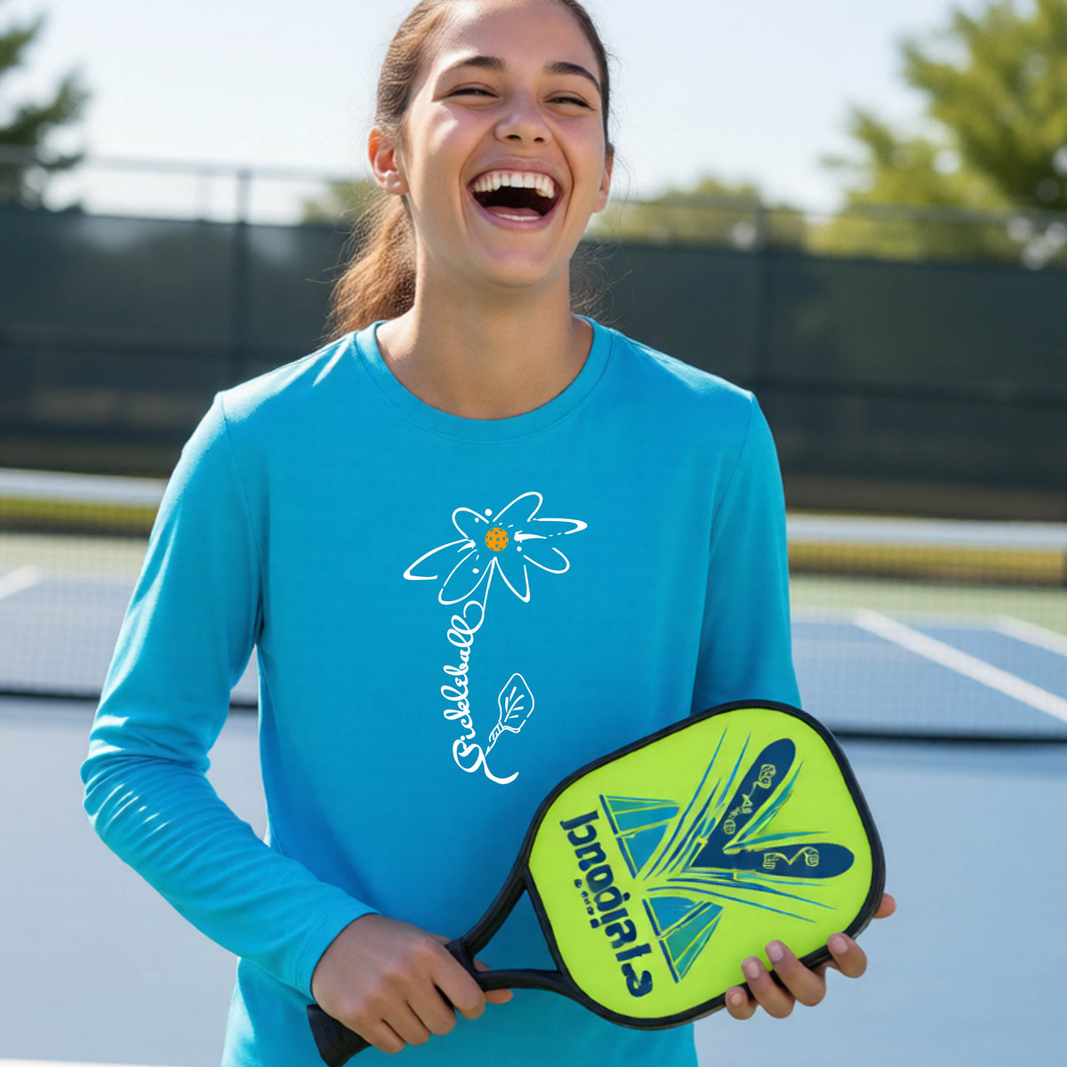 A youth wearing a bright blue long-sleeve t-shirt with a pickleball graphic of a flower with a colorful pickleball as the center with  the stem made of the word ‘PICKLEBALL’ and the leaf is a pickleball paddle, standing on a pickleball court.