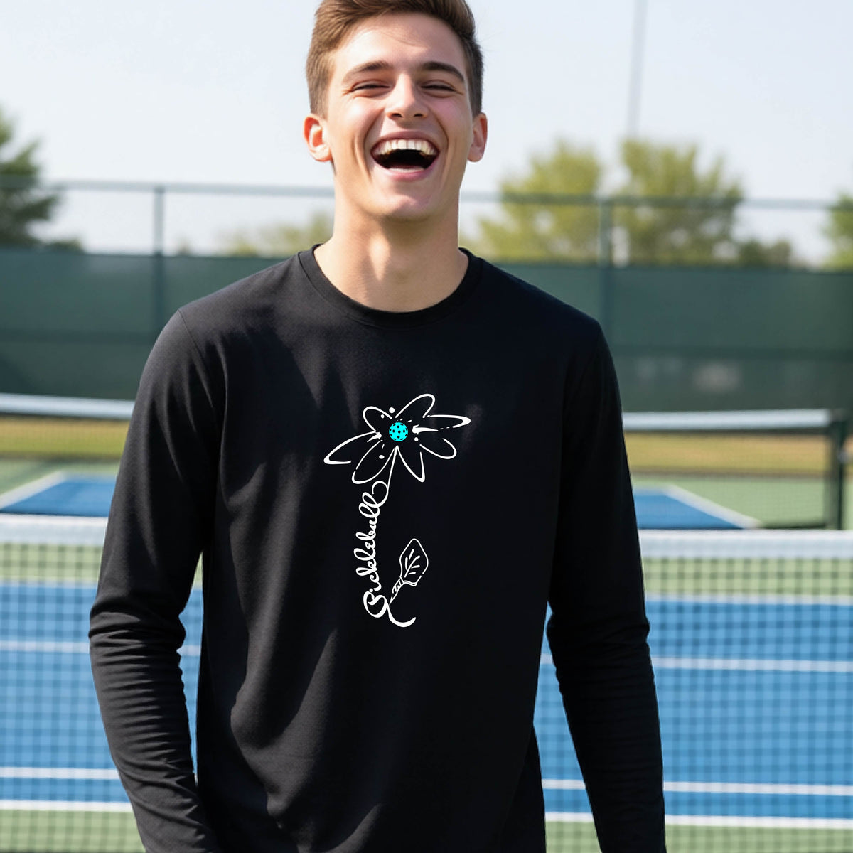 A youth wearing a black long-sleeve t-shirt with a pickleball graphic of a flower with a colorful pickleball as the center with  the stem made of the word ‘PICKLEBALL’ and the leaf is a pickleball paddle, standing on a pickleball court.