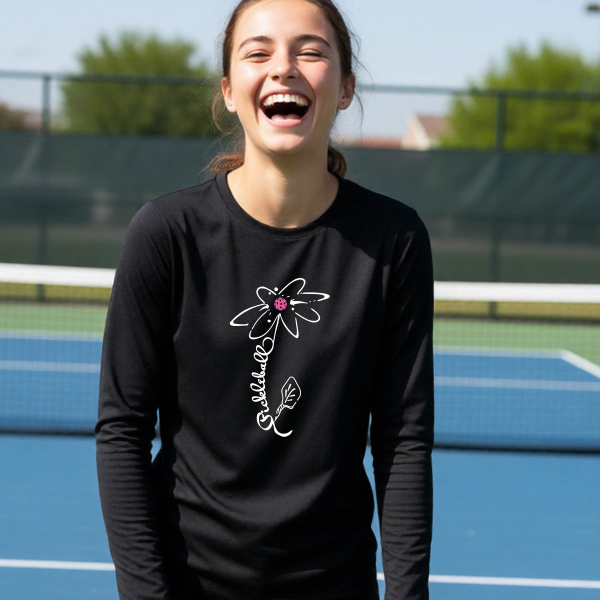 A youth wearing a black long-sleeve t-shirt with a pickleball graphic of a flower with a colorful pickleball as the center with  the stem made of the word ‘PICKLEBALL’ and the leaf is a pickleball paddle, standing on a pickleball court.