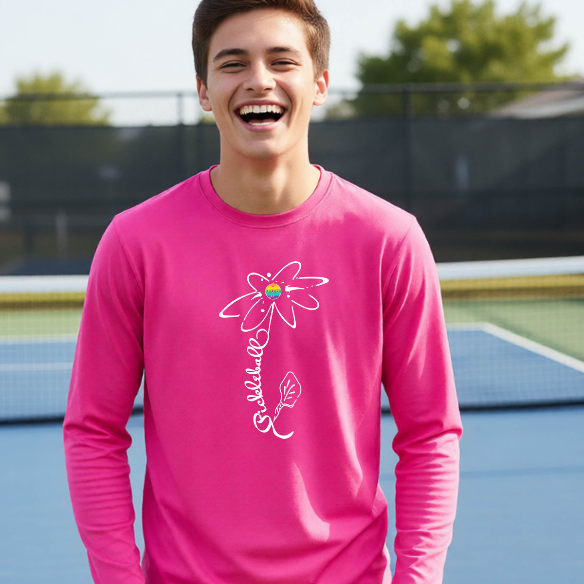 A youth wearing a pink long-sleeve t-shirt with a pickleball graphic of a flower with a colorful pickleball as the center with  the stem made of the word ‘PICKLEBALL’ and the leaf is a pickleball paddle, standing on a pickleball court.