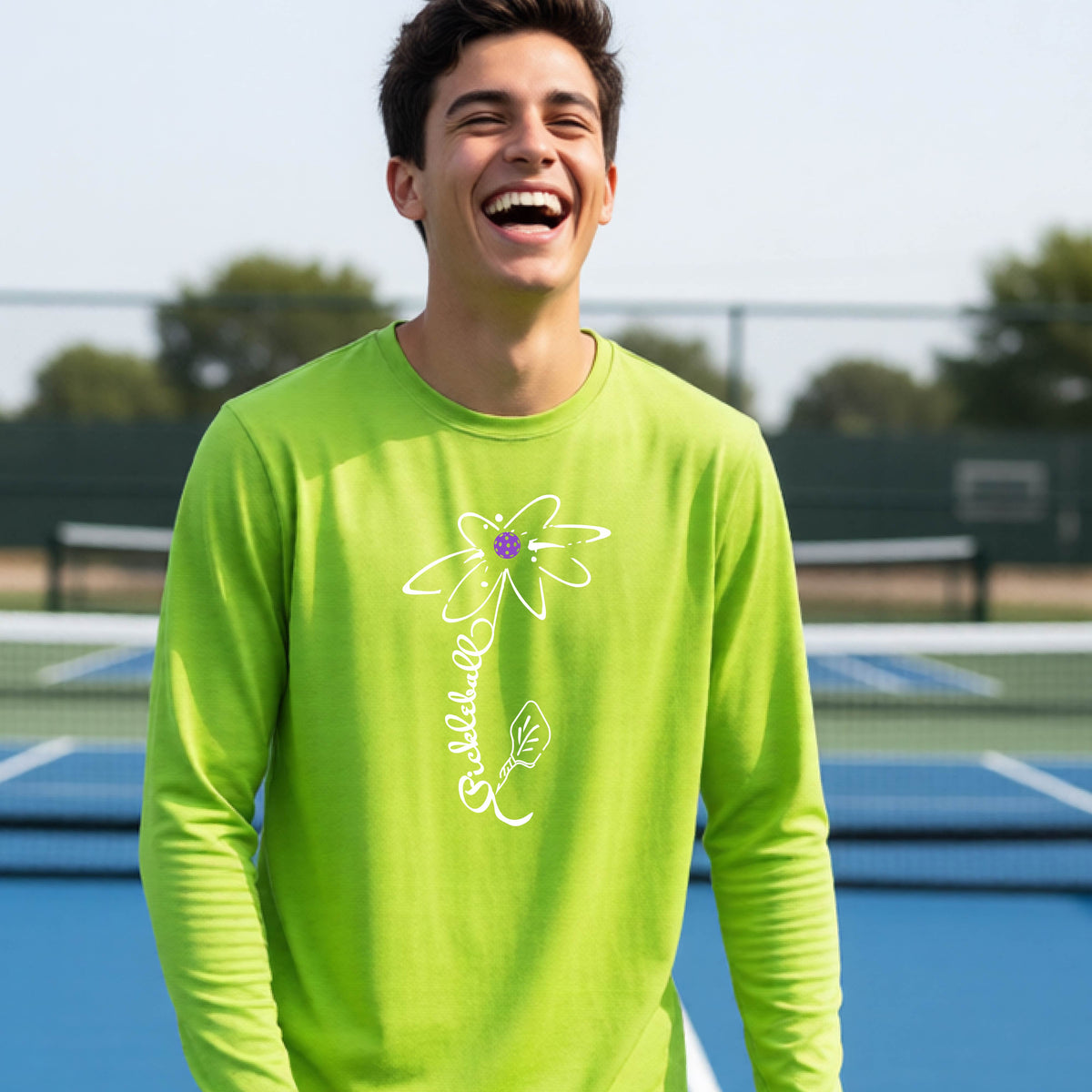 A youth wearing a bright lime green long-sleeve t-shirt with a pickleball graphic of a flower with a colorful pickleball as the center with  the stem made of the word ‘PICKLEBALL’ and the leaf is a pickleball paddle, standing on a pickleball court.