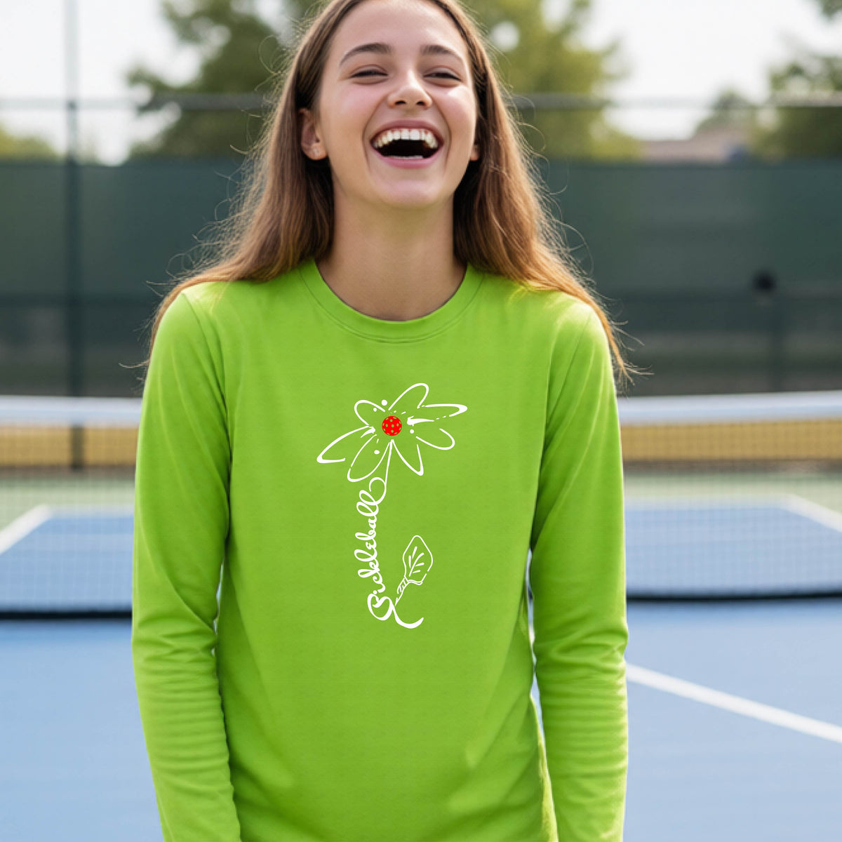 A youth wearing a lime green long-sleeve t-shirt with a pickleball graphic of a flower with a colorful pickleball as the center with  the stem made of the word ‘PICKLEBALL’ and the leaf is a pickleball paddle, standing on a pickleball court.