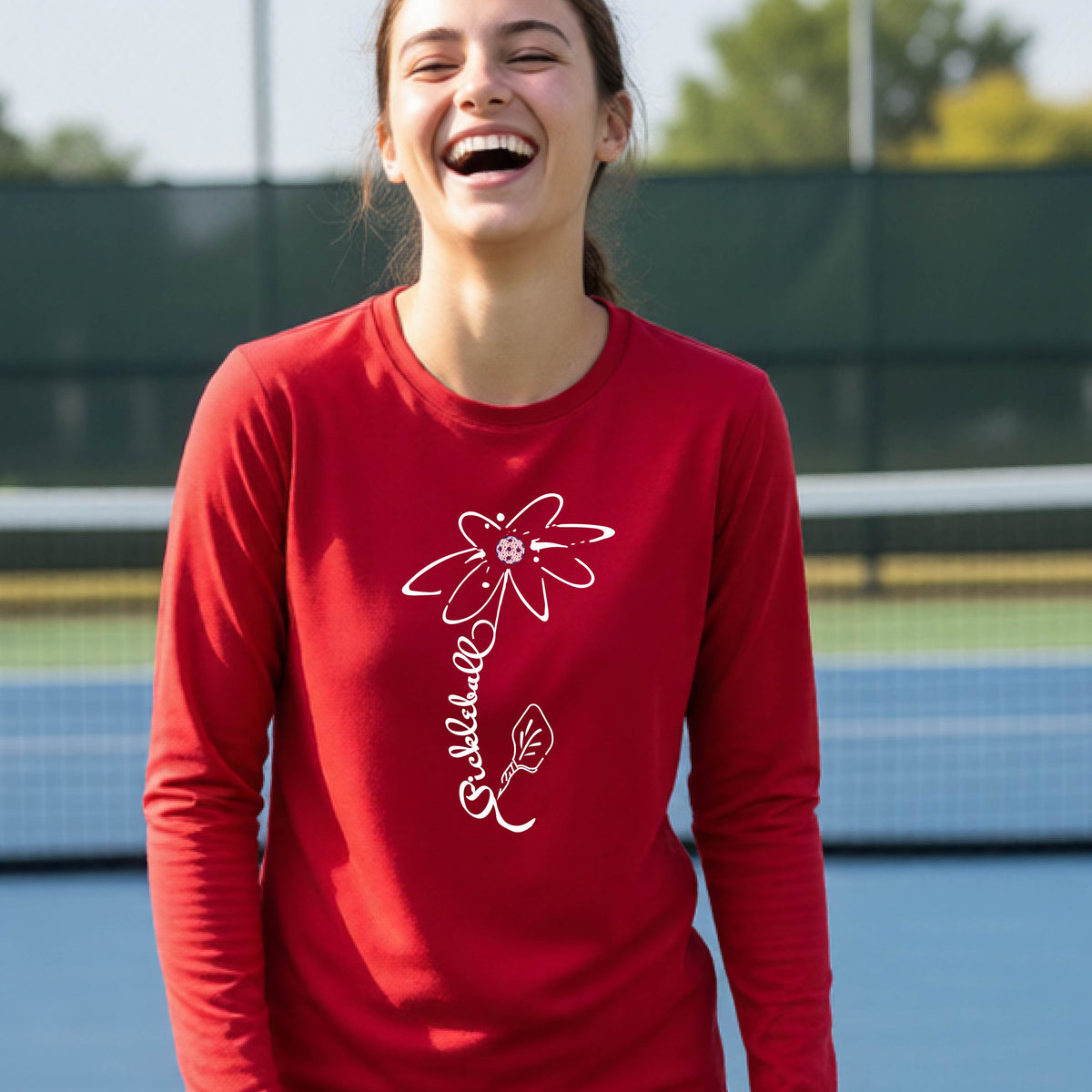 A youth wearing a red long-sleeve t-shirt with a pickleball graphic of a flower with a colorful pickleball as the center with  the stem made of the word ‘PICKLEBALL’ and the leaf is a pickleball paddle, standing on a pickleball court.