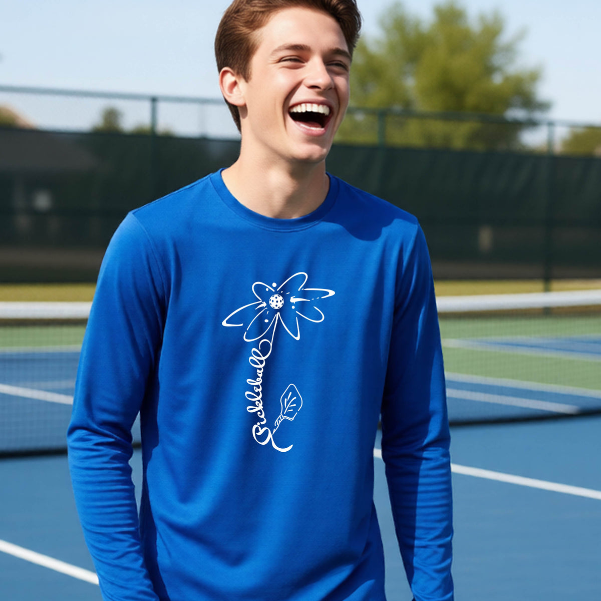 A youth wearing a royal blue long-sleeve t-shirt with a pickleball graphic of a flower with a colorful pickleball as the center with  the stem made of the word ‘PICKLEBALL’ and the leaf is a pickleball paddle, standing on a pickleball court.