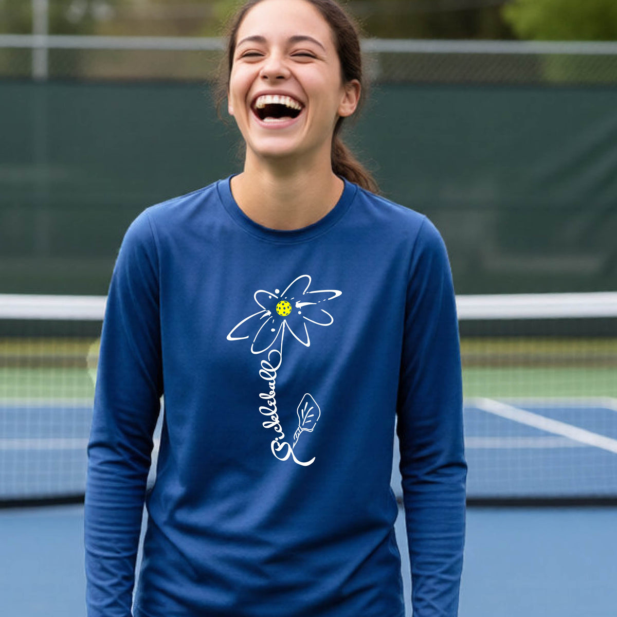 A youth wearing a royal blue long-sleeve t-shirt with a pickleball graphic of a flower with a colorful pickleball as the center with  the stem made of the word ‘PICKLEBALL’ and the leaf is a pickleball paddle, standing on a pickleball court.