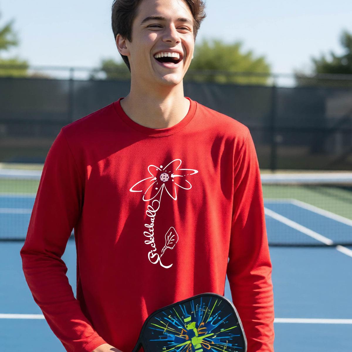 A youth wearing a red long-sleeve t-shirt with a pickleball graphic of a flower with a colorful pickleball as the center with  the stem made of the word ‘PICKLEBALL’ and the leaf is a pickleball paddle, standing on a pickleball court.