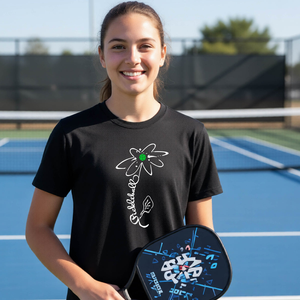 A teenager wearing a black short-sleeve  t-shirt with a pickleball graphic of a flower with a flower that has a colorful pickleball as the center and the stem is made of the word ‘PICKLEBALL’ and the leaf is a pickleball paddle, standing on a pickleball court.