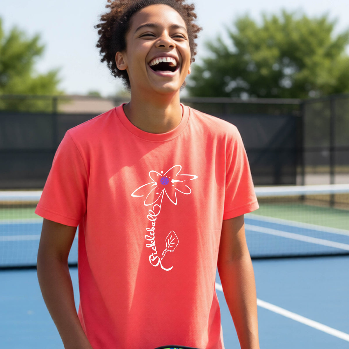 A teenager wearing a coral short-sleeve  t-shirt with a pickleball graphic of a flower with a flower that has a colorful pickleball as the center and the stem is made of the word ‘PICKLEBALL’ and the leaf is a pickleball paddle, standing on a pickleball court.