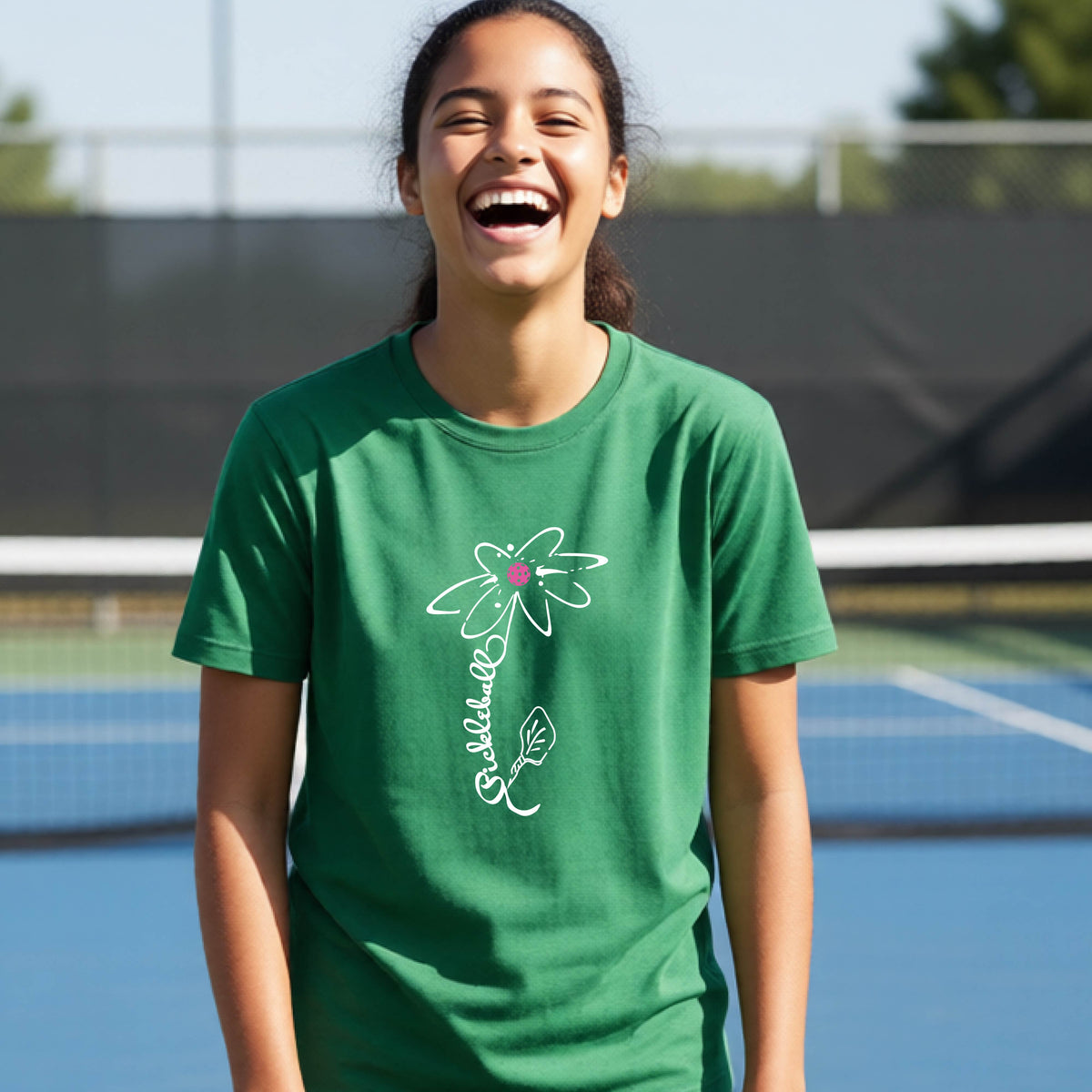 A teenager wearing a green short-sleeve  t-shirt with a pickleball graphic of a flower with a flower that has a colorful pickleball as the center and the stem is made of the word ‘PICKLEBALL’ and the leaf is a pickleball paddle, standing on a pickleball court.