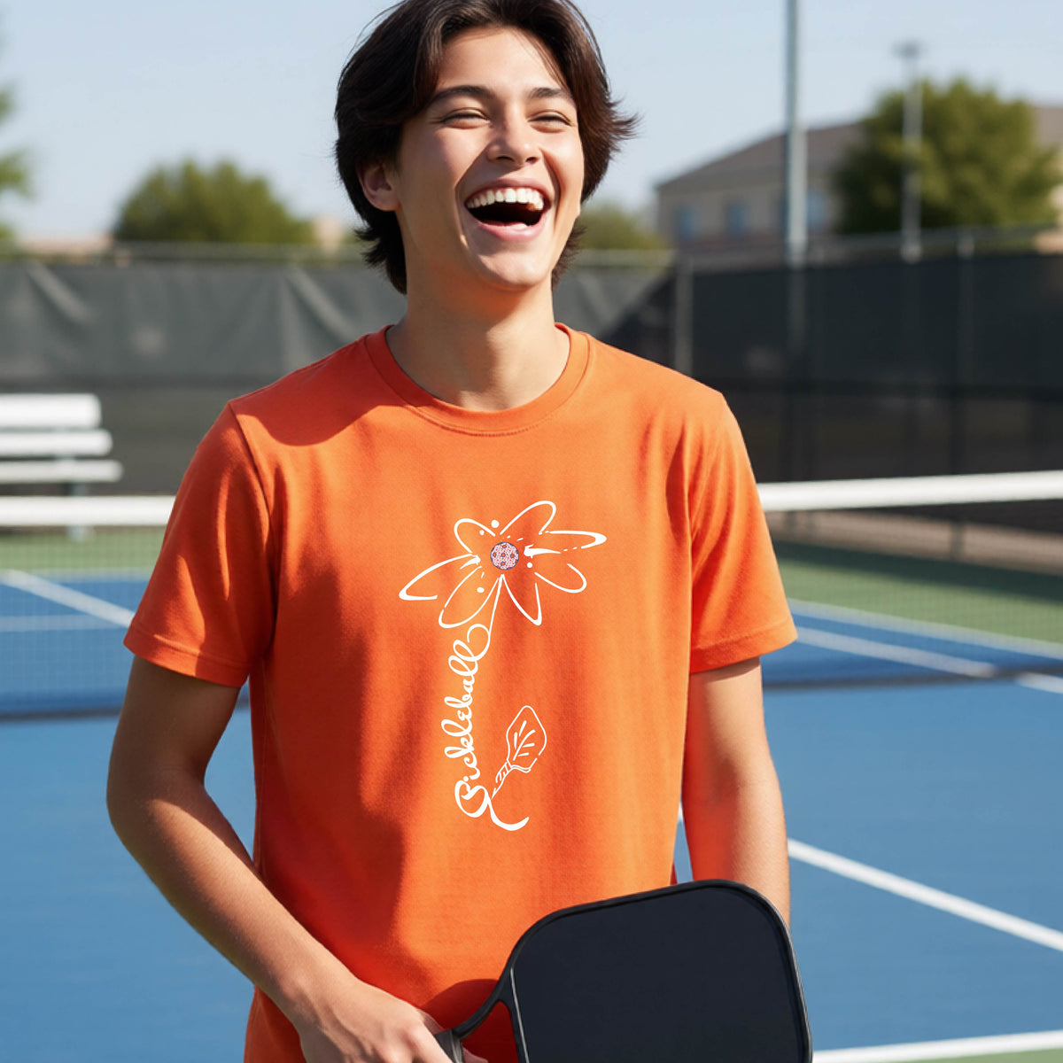 A teenager wearing an orange short-sleeve  t-shirt with a pickleball graphic of a flower with a flower that has a colorful pickleball as the center and the stem is made of the word ‘PICKLEBALL’ and the leaf is a pickleball paddle, standing on a pickleball court.