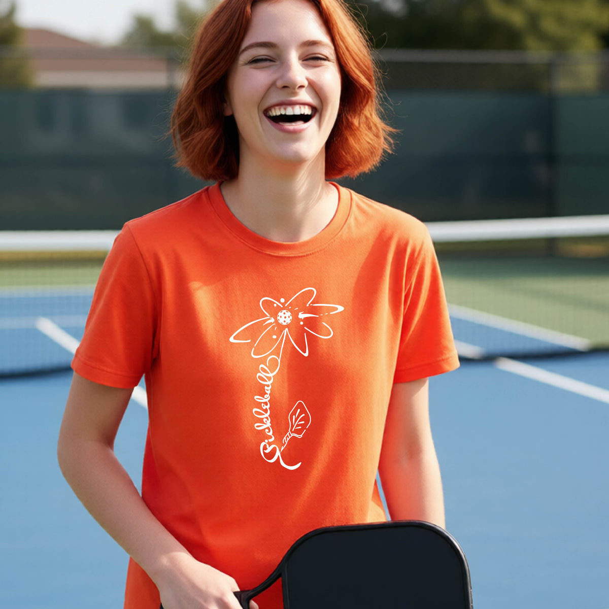 A teenager wearing an orange short-sleeve  t-shirt with a pickleball graphic of a flower with a flower that has a colorful pickleball as the center and the stem is made of the word ‘PICKLEBALL’ and the leaf is a pickleball paddle, standing on a pickleball court.
