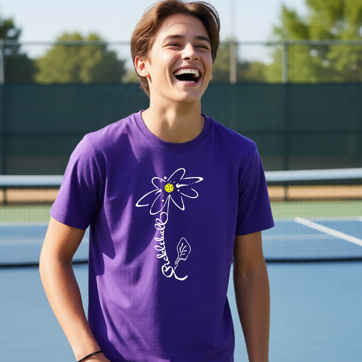 A teenager wearing a purple short-sleeve  t-shirt with a pickleball graphic of a flower with a flower that has a colorful pickleball as the center and the stem is made of the word ‘PICKLEBALL’ and the leaf is a pickleball paddle, standing on a pickleball court.