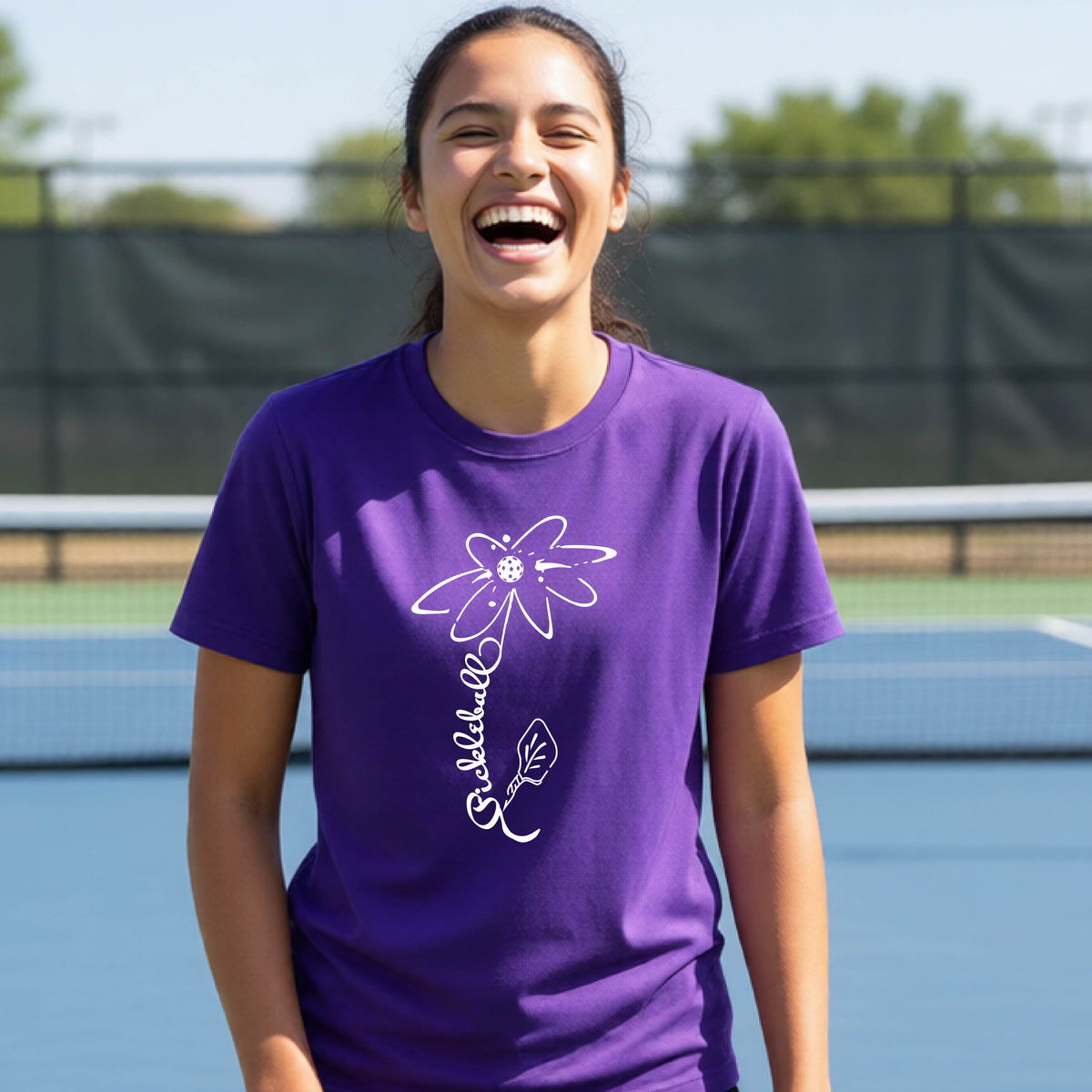 A teenager wearing a purple short-sleeve  t-shirt with a pickleball graphic of a flower with a flower that has a colorful pickleball as the center and the stem is made of the word ‘PICKLEBALL’ and the leaf is a pickleball paddle, standing on a pickleball court.