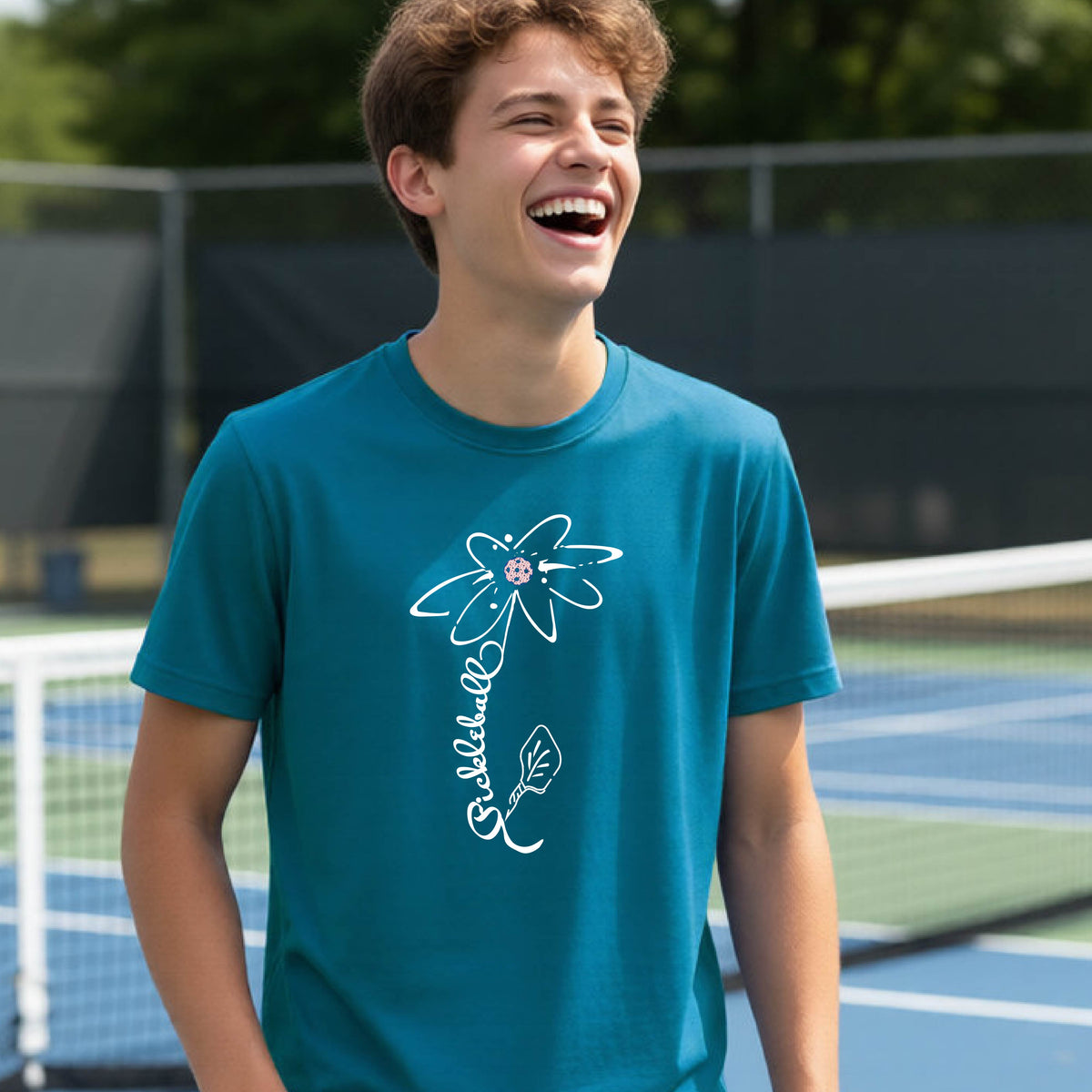A teenager wearing a teal short-sleeve  t-shirt with a pickleball graphic of a flower with a flower that has a colorful pickleball as the center and the stem is made of the word ‘PICKLEBALL’ and the leaf is a pickleball paddle, standing on a pickleball court.