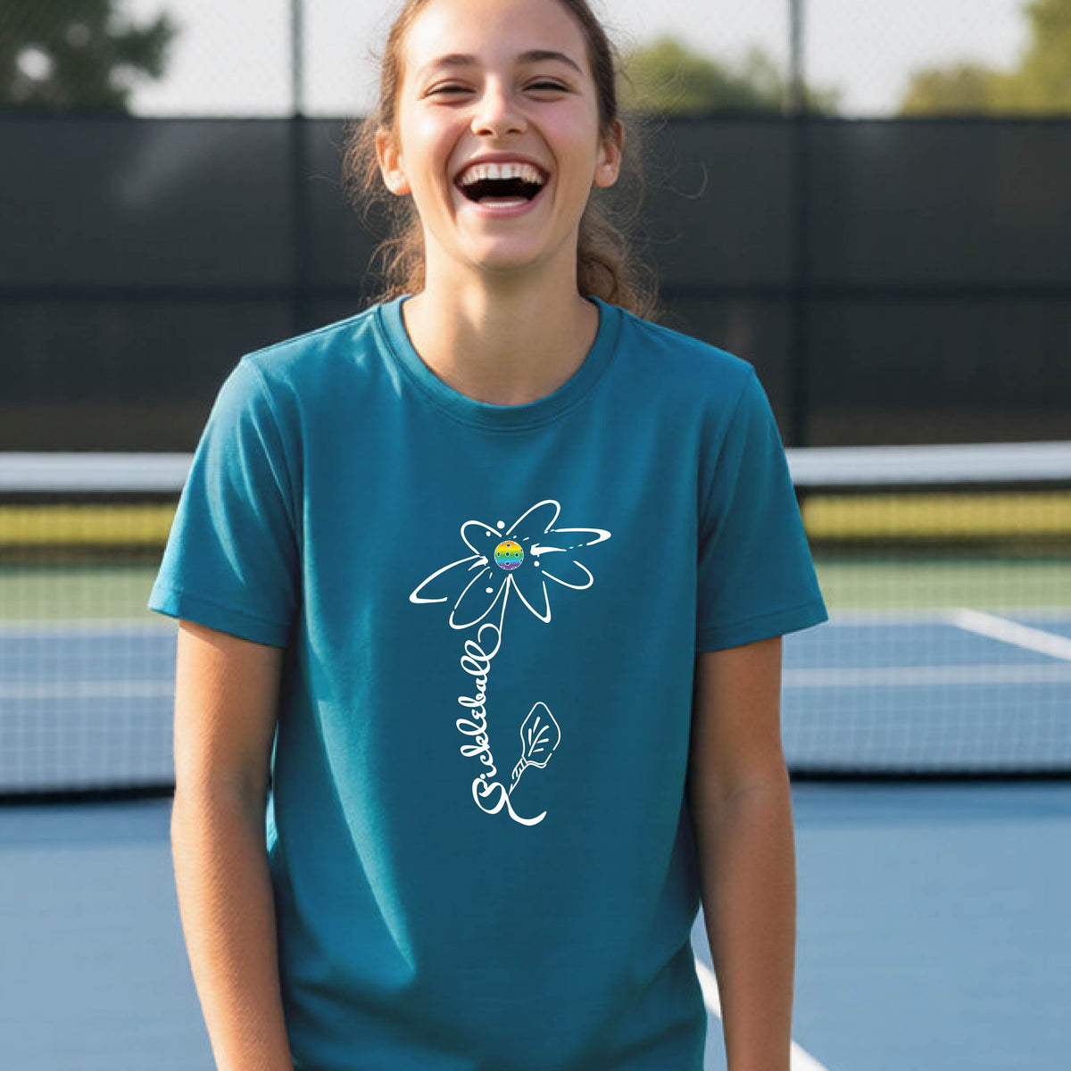 A teenager wearing a teal short-sleeve  t-shirt with a pickleball graphic of a flower with a flower that has a colorful pickleball as the center and the stem is made of the word ‘PICKLEBALL’ and the leaf is a pickleball paddle, standing on a pickleball court.