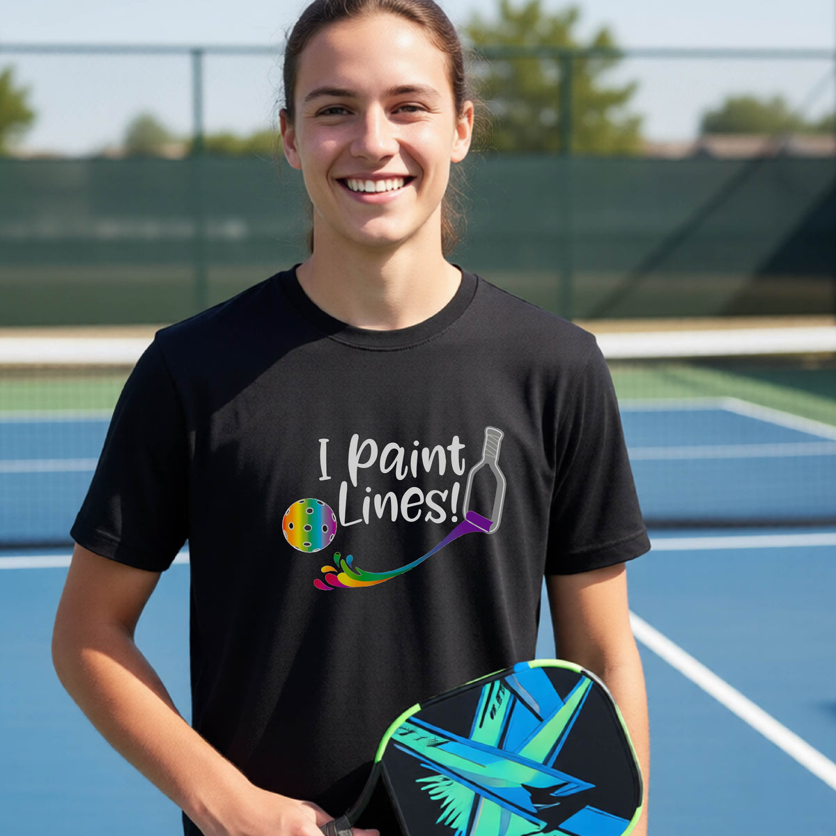 A young adult on an outdoor pickleball court wearing a black short-sleeve t-shirt with a graphic that reads 'THIS IS MY PICKLEBALL SHIRT' in white with a yellow pickleball graphic on the back.