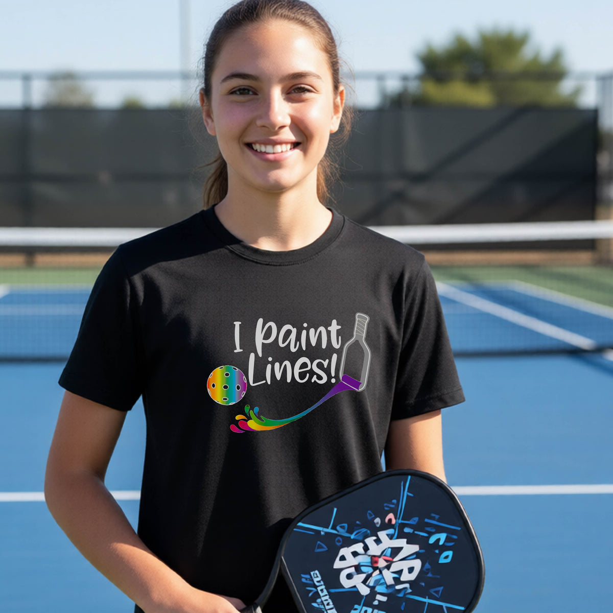 A young adult on an outdoor pickleball court wearing a black short-sleeve t-shirt with a graphic that reads 'THIS IS MY PICKLEBALL SHIRT' in white with a yellow pickleball graphic on the back.