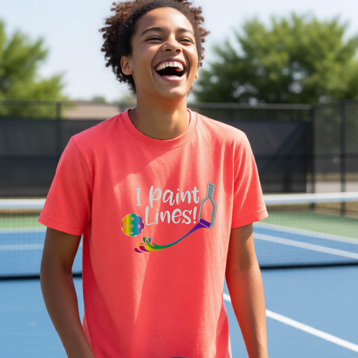 A young adult on an outdoor pickleball court wearing a coral pink short-sleeve t-shirt with a graphic that reads 'THIS IS MY PICKLEBALL SHIRT' in white with a yellow pickleball graphic on the back.