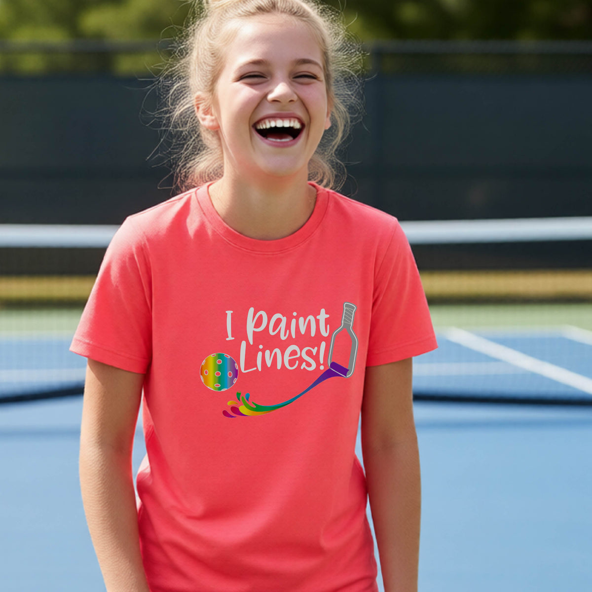A young adult on an outdoor pickleball court wearing a coral pink short-sleeve t-shirt with a graphic that reads 'THIS IS MY PICKLEBALL SHIRT' in white with a yellow pickleball graphic on the back.