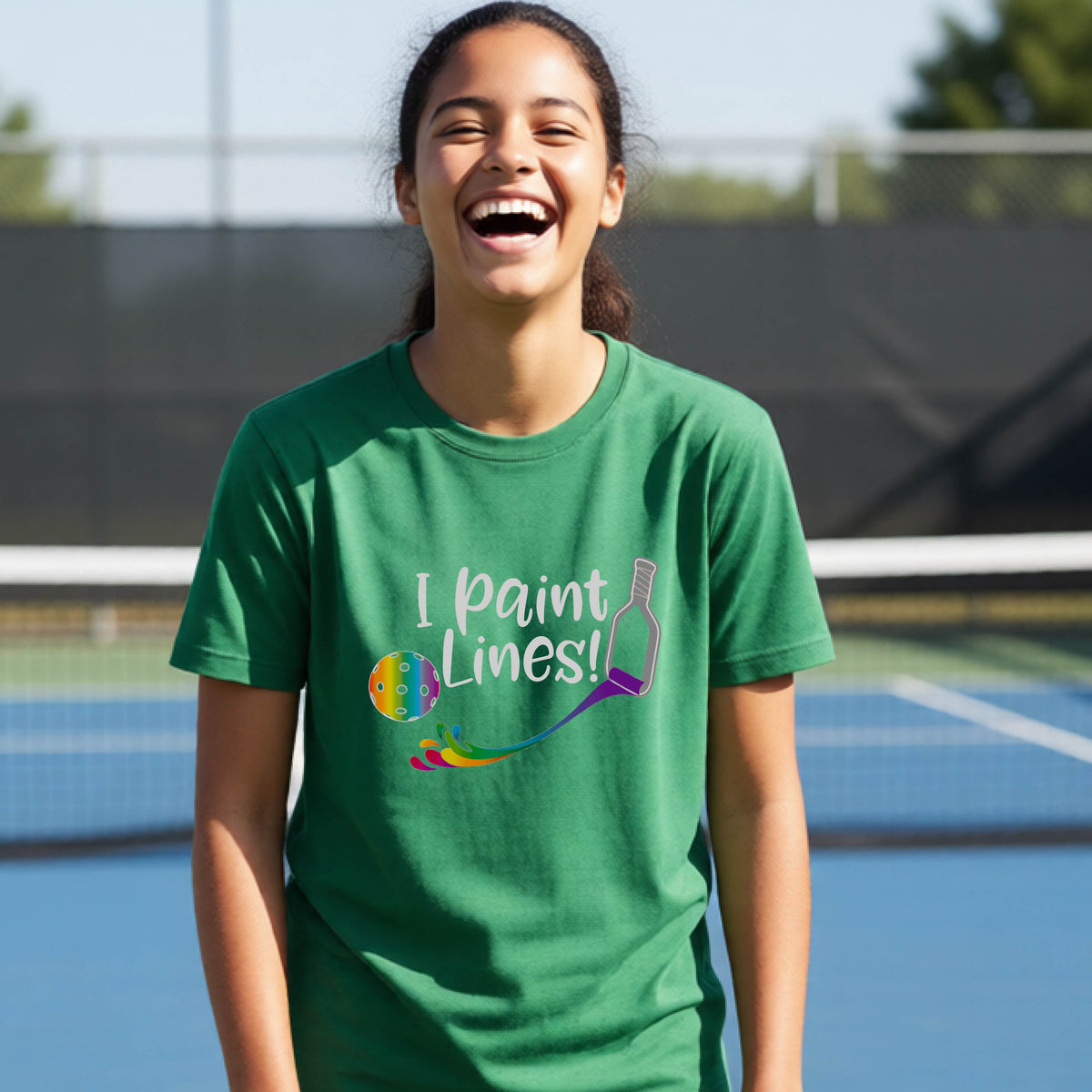 A young adult on an outdoor pickleball court wearing a green short-sleeve t-shirt with a graphic that reads 'THIS IS MY PICKLEBALL SHIRT' in white with a yellow pickleball graphic on the back.