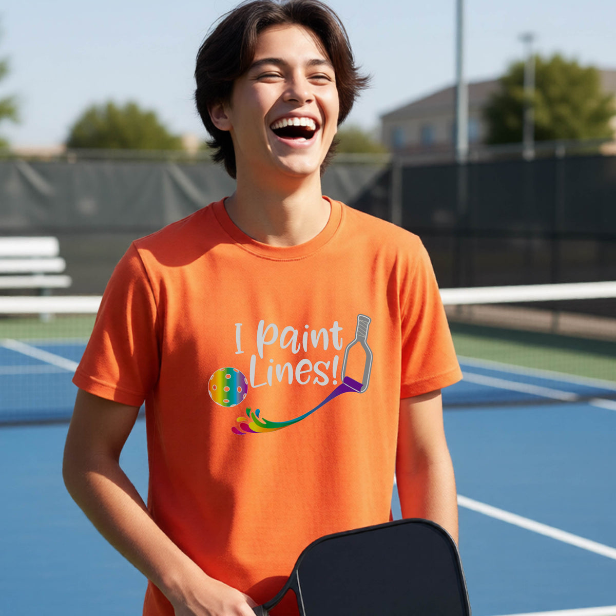 A young adult on an outdoor pickleball court wearing an orange short-sleeve t-shirt with a graphic that reads 'THIS IS MY PICKLEBALL SHIRT' in white with a yellow pickleball graphic on the back.