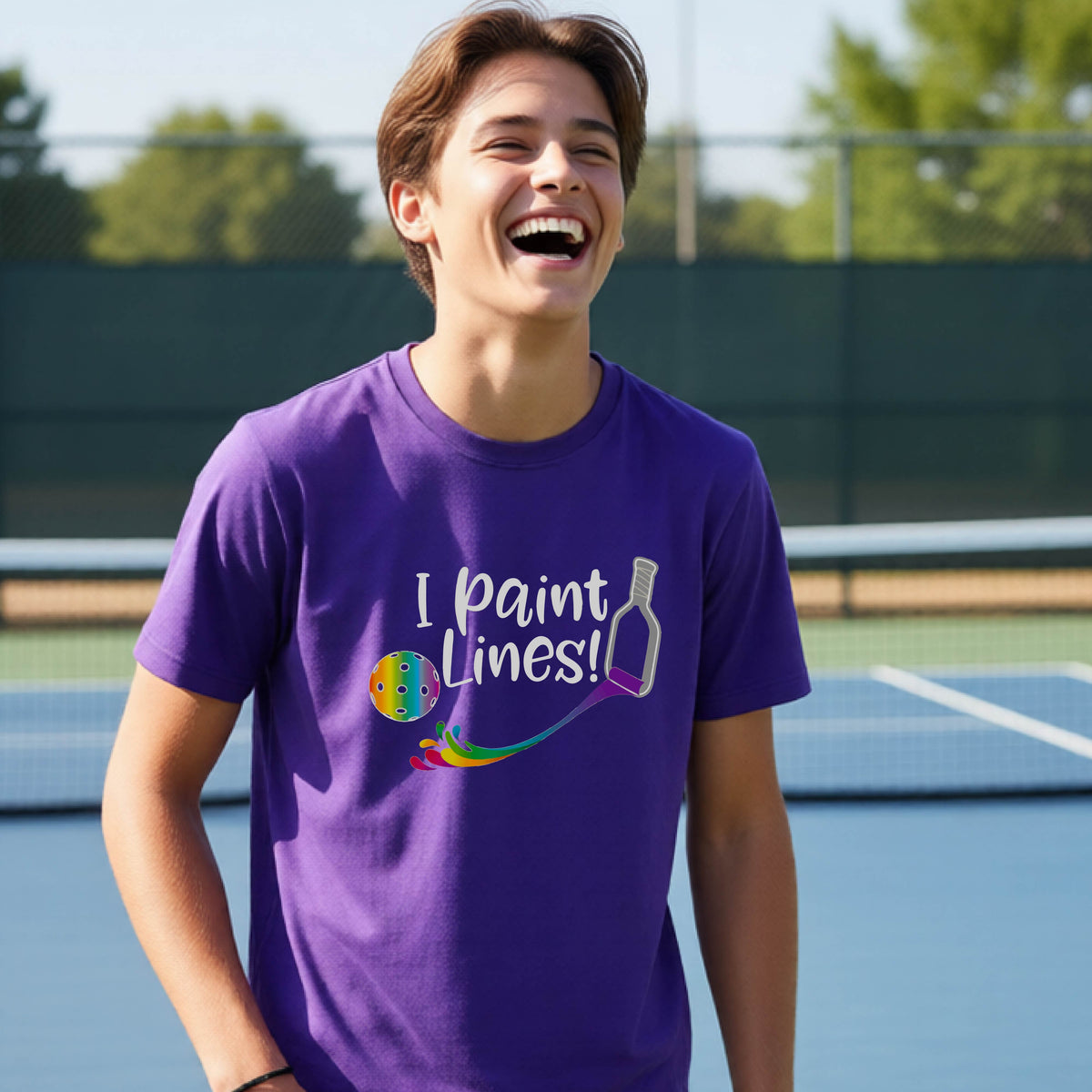 A young adult on an outdoor pickleball court wearing a purple short-sleeve t-shirt with a graphic that reads 'THIS IS MY PICKLEBALL SHIRT' in white with a yellow pickleball graphic on the back.
