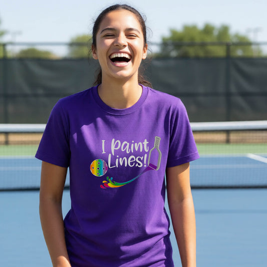 A young adult on an outdoor pickleball court wearing a purple short-sleeve t-shirt with a graphic that reads 'THIS IS MY PICKLEBALL SHIRT' in white with a yellow pickleball graphic on the back.