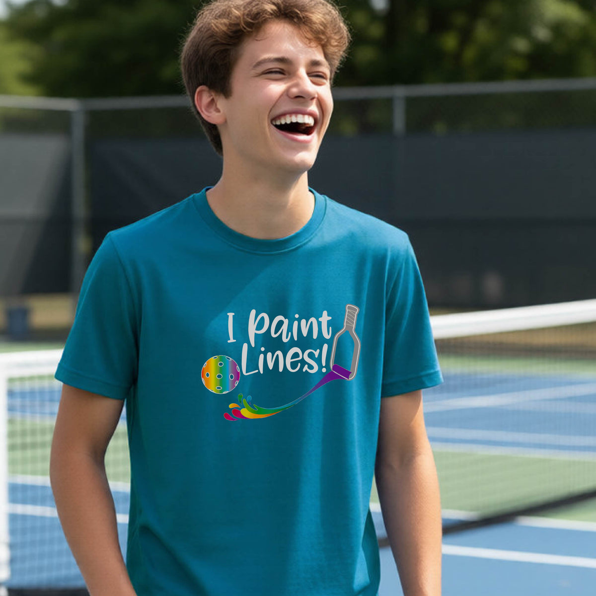 A young adult on an outdoor pickleball court wearing a teal short-sleeve t-shirt with a graphic that reads 'THIS IS MY PICKLEBALL SHIRT' in white with a yellow pickleball graphic on the back.